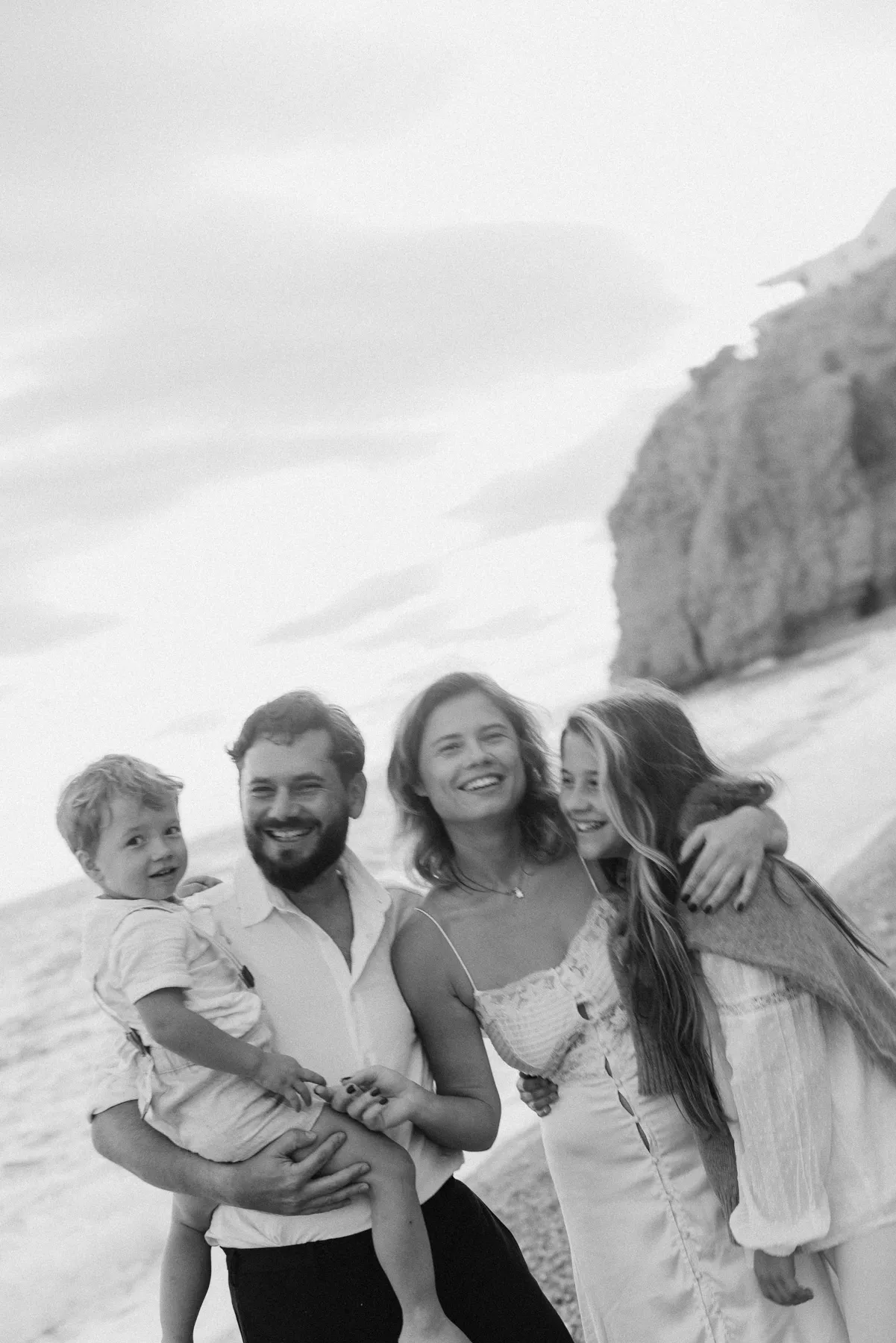 Family with two children laughing on the beach in front of rocks, black and white