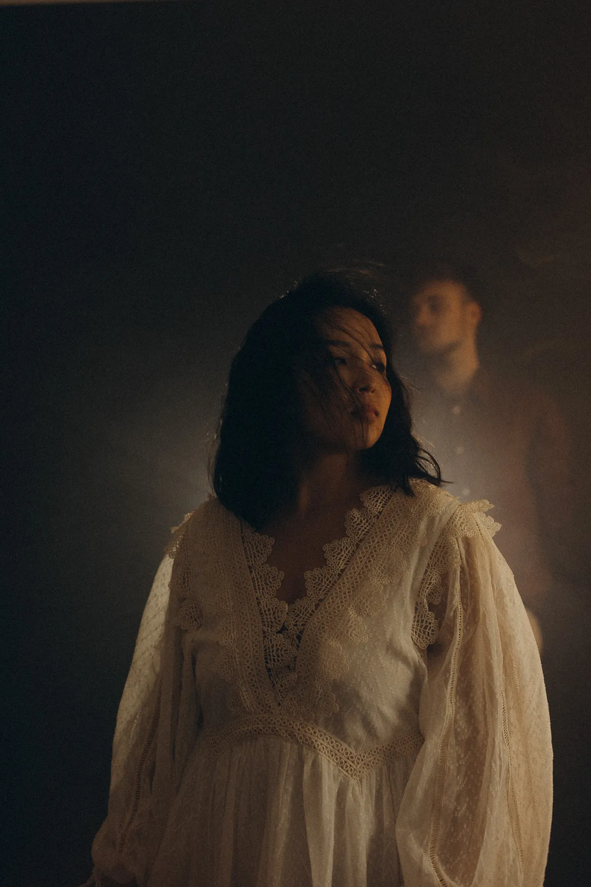 Woman in a light dress in a dark room, her partner in the background