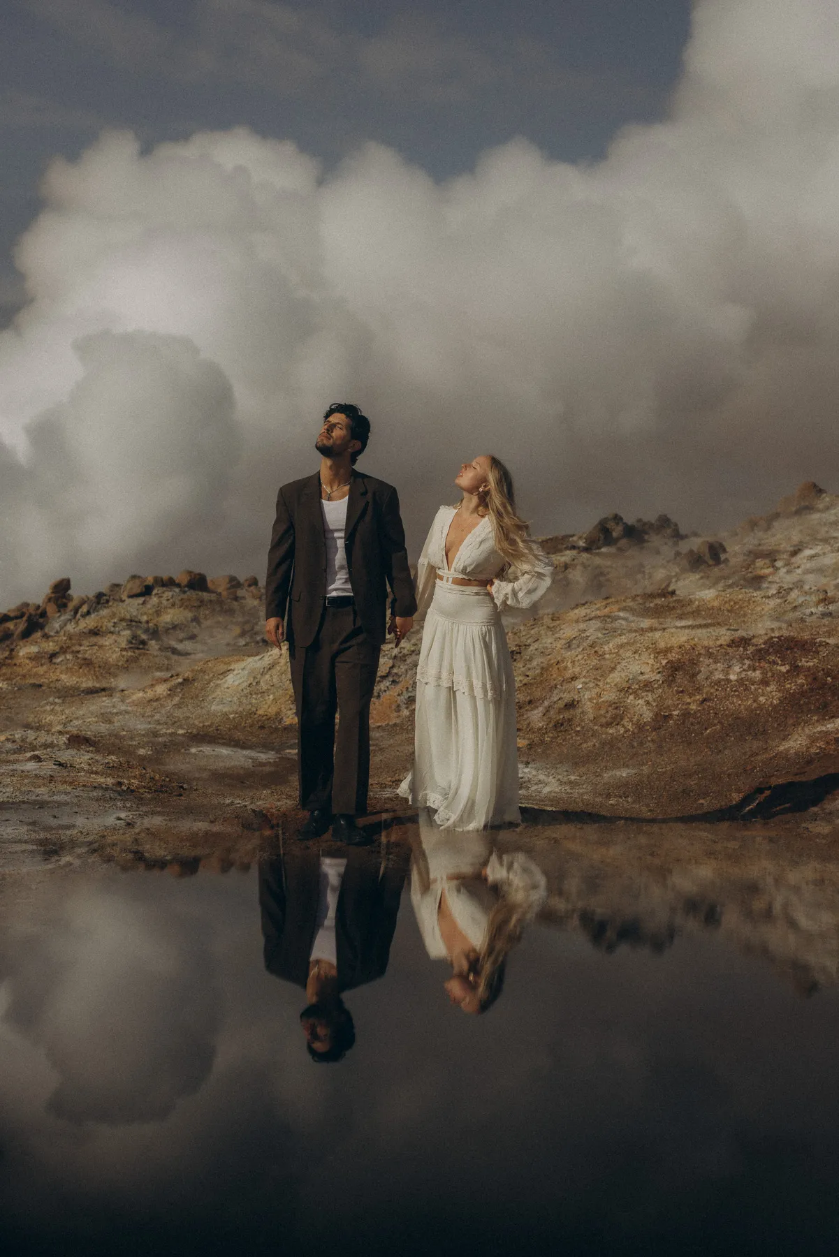Paar in Hochzeitskleidung vor dampfenden Quellen auf Island, gespiegelt im Wasser