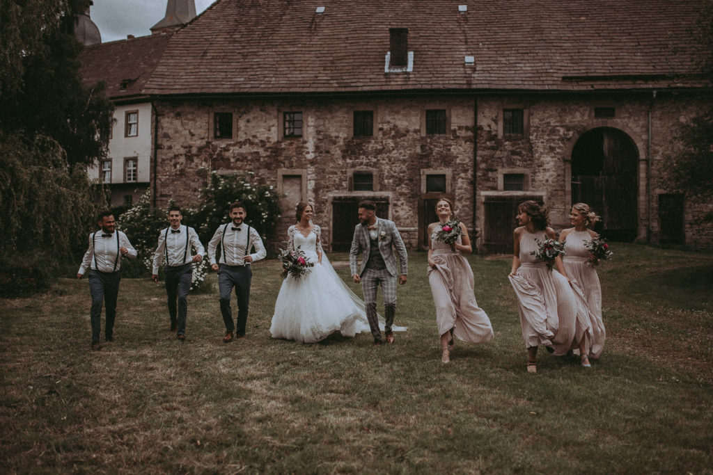 Foto von Braut und Bräutigam mit Trauzeugen und Brautjungfern auf einer Wiese vor einem alten Farmhaus.