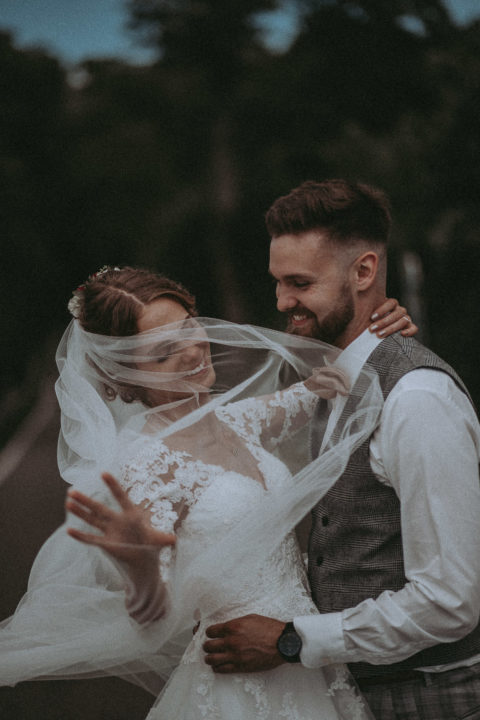 lachendes Brautpaar tanzend auf einer Wiese, Braut wirbelt Schleier